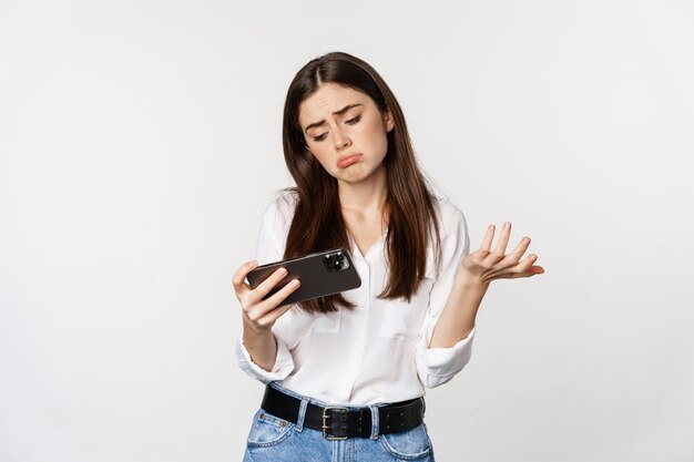 Sad woman losing in mobile video game, looking upset and disappointed at smartphone, sulking, standing over white background