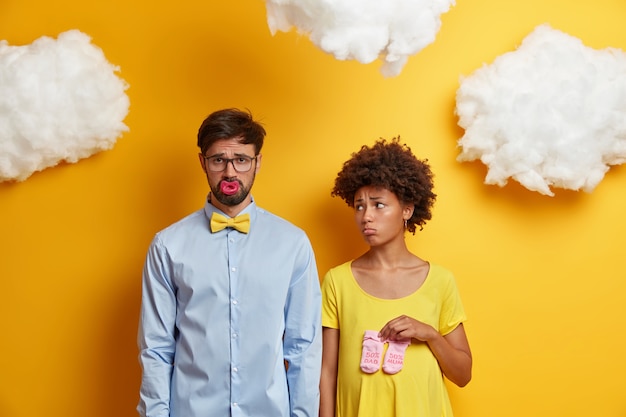 Free photo sad woman and man expect baby, find out bad news, pose with nipple and childs socks, get ready for becoming parents, stand next to each other against yellow . young family prepare for birth