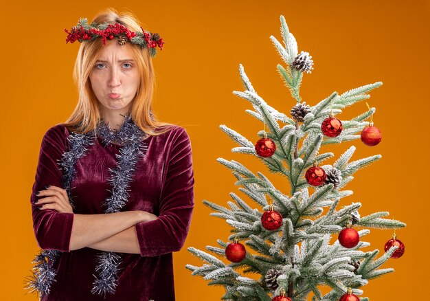 Sad young beautiful girl standing nearby christmas tree wearing red dress and wreath with garland on neck crossing hands isolated on orange background