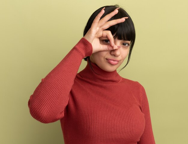 Sad young brunette caucasian girl through fingers isolated on olive green wall with copy space