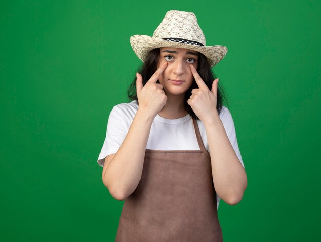 Sad young brunette female gardener in optical glasses and uniform wearing gardening hat pulls down eyelids isolated on green wall