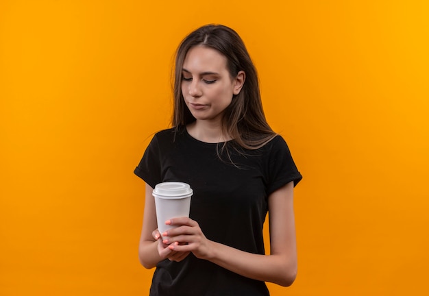 Sad young caucasian girl wearing black t-shirt looking at cup of coffee on isolated orange background