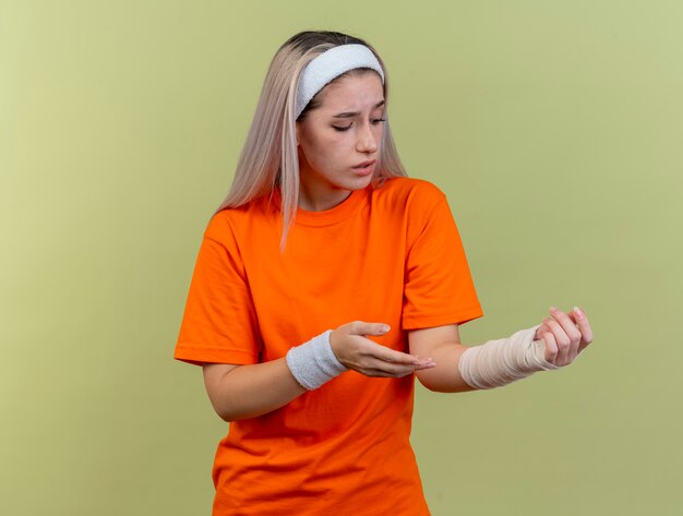 Sad young caucasian sporty girl with braces wearing headband and wristbands points and looks at hand 