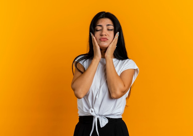 Sad young caucasian woman puts hands on face standing with closed eyes