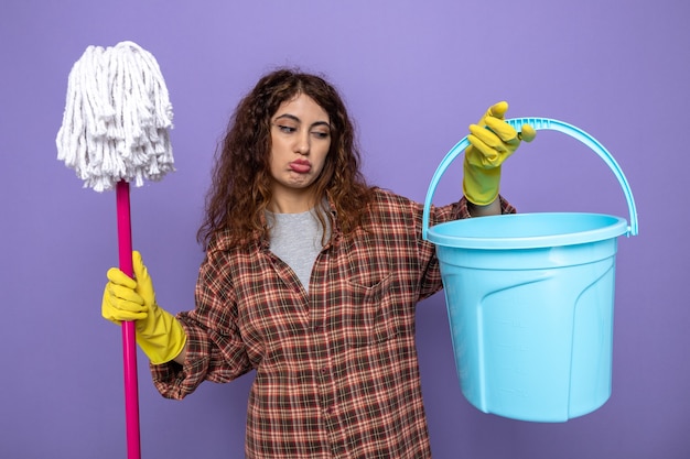 Free photo sad young cleaning woman wearing gloves holding mop looking at bucket in her hand