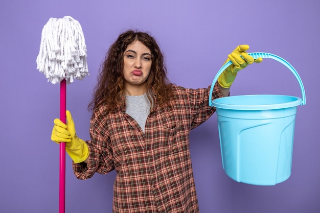 Free Photo sad young cleaning woman wearing gloves holding mop with bucket isolated on purple wall