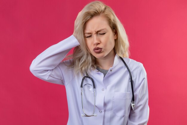 Sad young doctor wearing stethoscope in medical gown put her hand on aching head on red backgroung