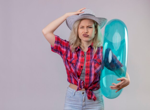 Sad young female traveler wearing red shirt holding inflatable ring put her hand on hat on isolated white wall