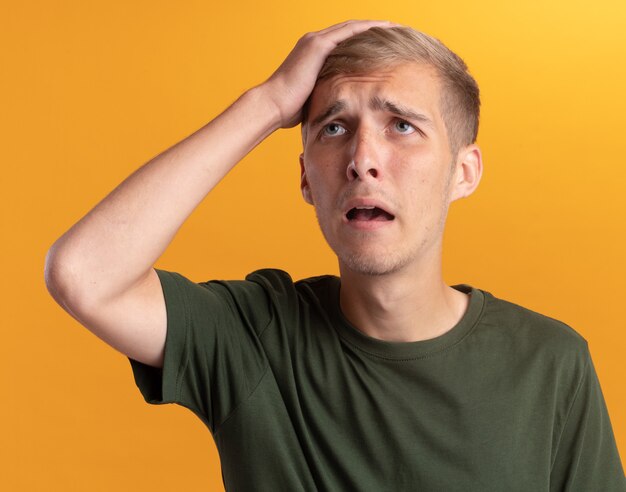 Sad young handsome guy wearing green shirt putting hand on head isolated on yellow wall