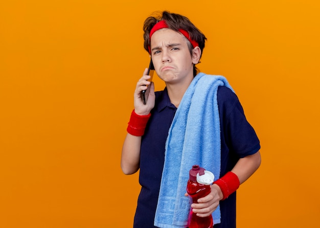 Sad young handsome sporty boy with towel on shoulder, holding water bottle and talking on phone