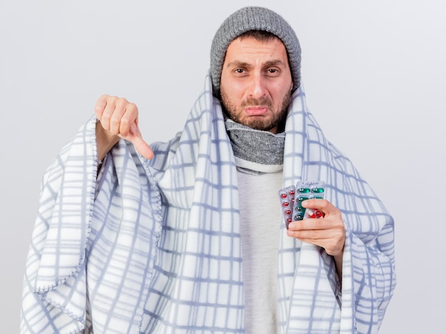 Sad young ill man wearing winter hat and scarf wrapped in plaid holding pills