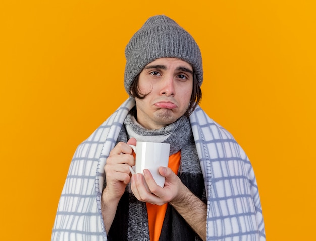 Free photo sad young ill man wearing winter hat with scarf wrapped in plaid holding cup of tea isolated on orange