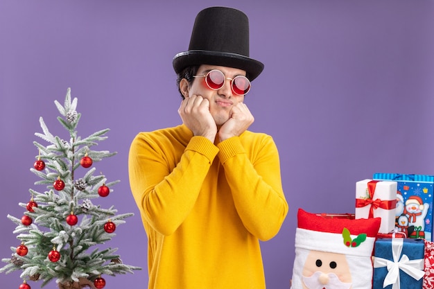 Free photo sad young man in yellow turtleneck and glasses wearing black hat standing next to a christmas tree and presents over purple background