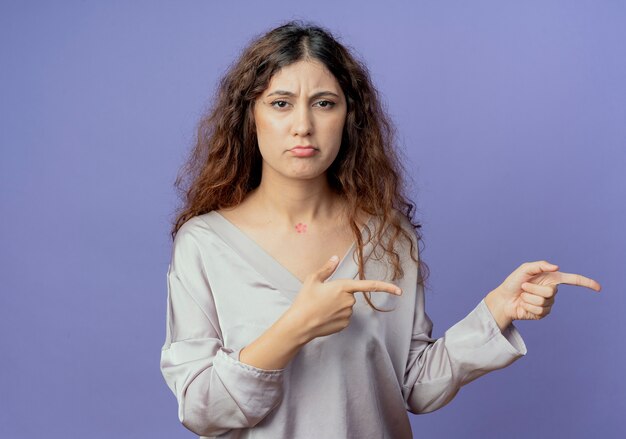 Sad young pretty girl points at side isolated on blue wall with copy space