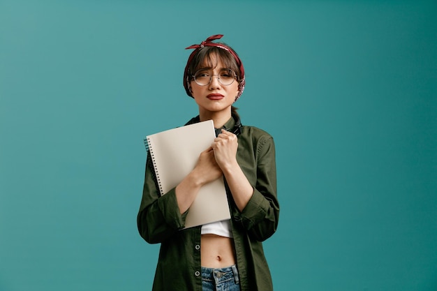 Free photo sad young student girl wearing bandana glasses and headphones around neck holding large note pad hugging it keeping hands together looking at camera isolated on blue background