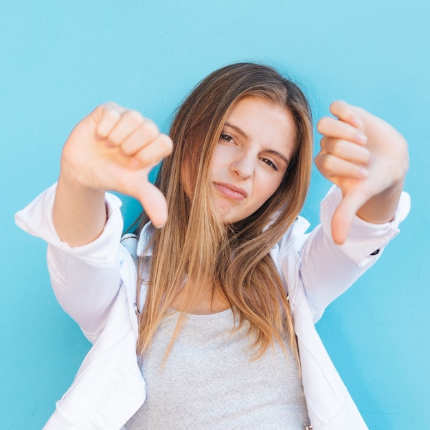 Sad young woman showing thumbs down in front of camera against blue background