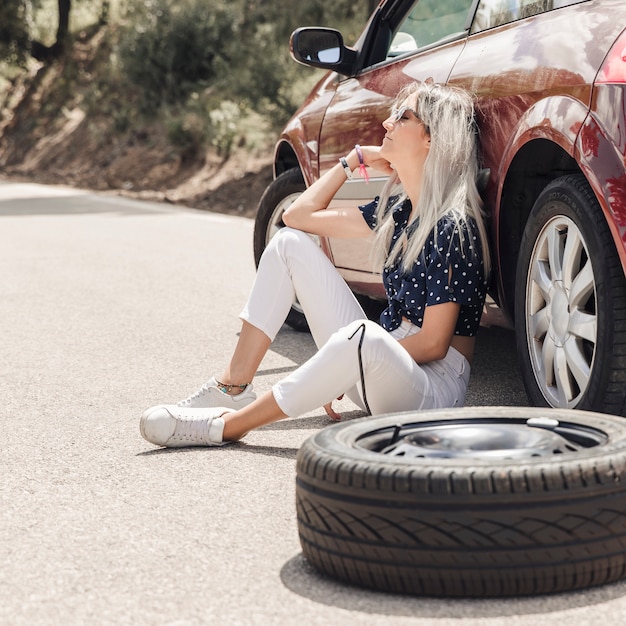 Sad young woman sitting near the broken down car on road