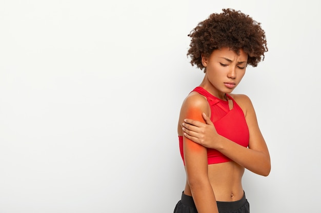 Free photo sad young woman suffers from painful feeling in arm muscle, stands sideways against white background, injured shoulder during workout session