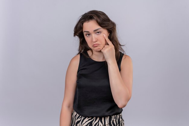 Sad young woman wearing black undershirt points eye on white wall