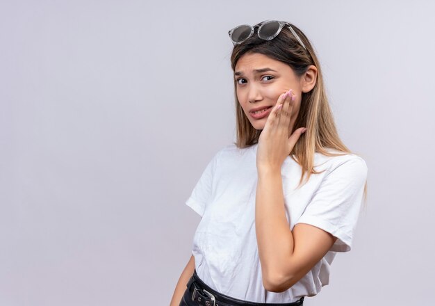 A sad young woman in white t-shirt in sunglasses keeping hand on cheek and having ache while looking on a white wall