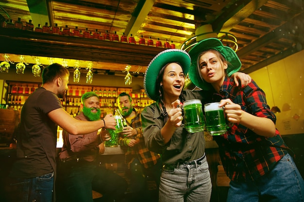 Free photo saint patrick's day party. happy friends is celebrating and drinking green beer. young men and women wearing a green hats. pub interior.