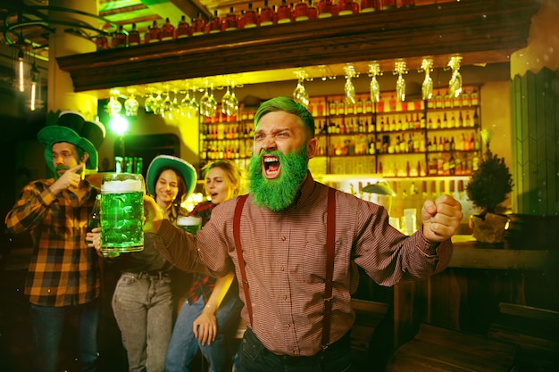 Saint Patrick's Day Party. Happy friends is celebrating and drinking green beer. Young men and women wearing a green hats. Pub Interior.