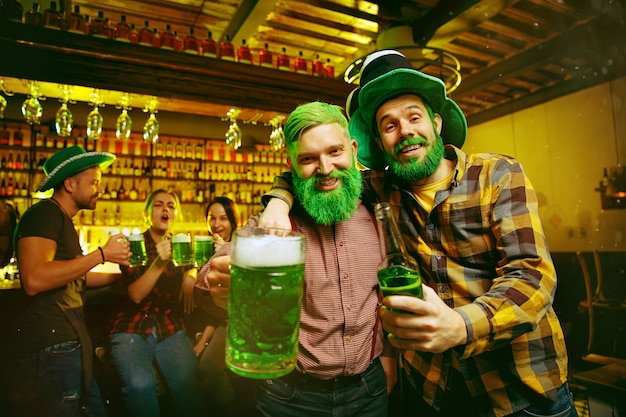 Saint Patrick's Day Party. Happy friends is celebrating and drinking green beer. Young men and women wearing a green hats. Pub Interior.