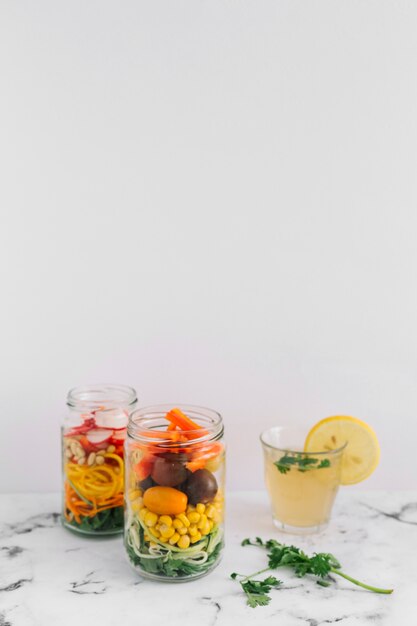 Salad vegetables in two mason jar with glass of lemon juice on marble top