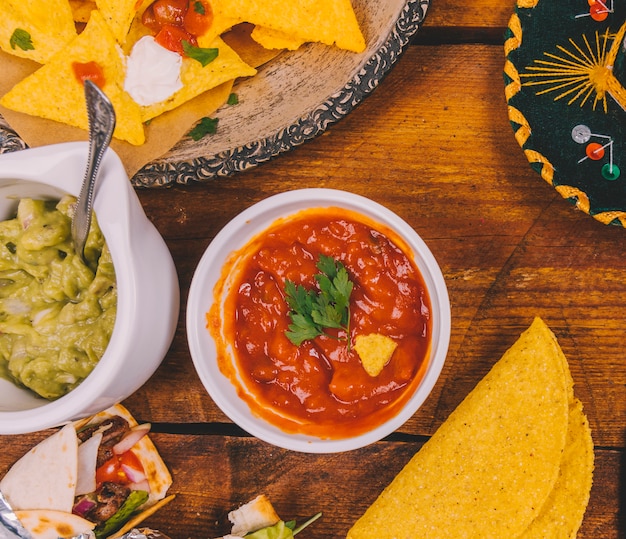 Salsa sauce; guacamole; tasty mexican nachos; tortilla and wrap mexican tacos on wooden table