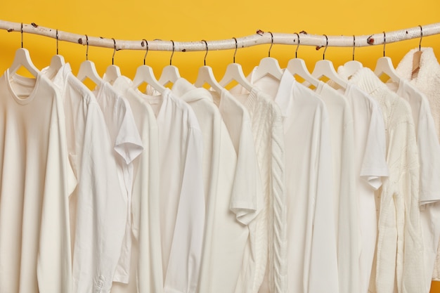 Free photo same white clothes on wooden racks in closet. collection of clothing on hangers, isolated over yellow background.