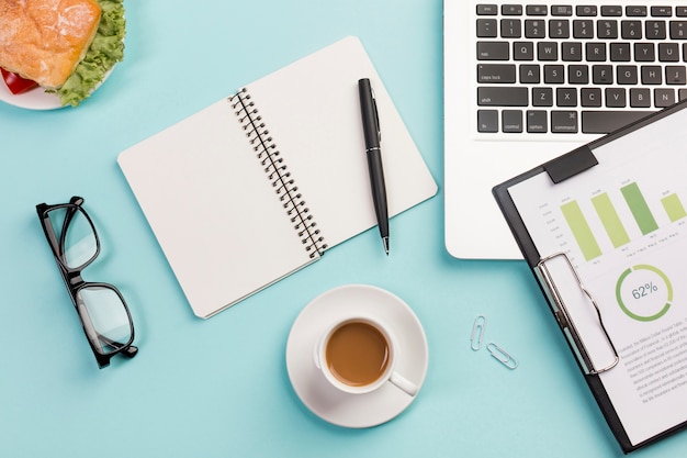 Free photo sandwich,coffee cup,eyeglasses,spiral notepad,pen,laptop and clipboard with budget plan on blue desk