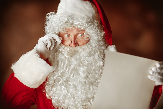 Free photo santa claus with a luxurious white beard, santa's hat and a red costume reading letter