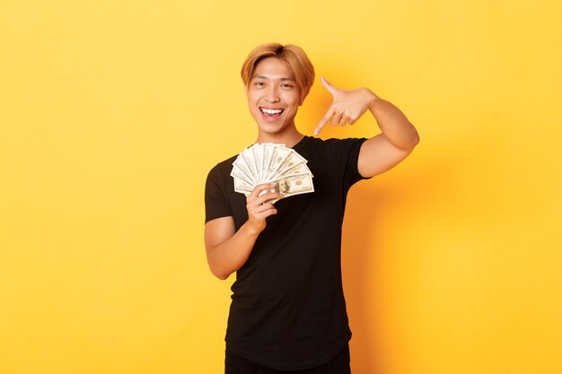 Sassy good-looking korean blond guy, smiling happy and pointing finger at money, winning cash, standing yellow wall