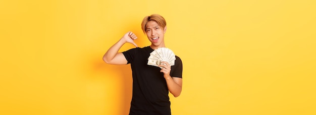 Sassy handsome asian guy pointing finger at cash and looking pleased korean man borrowed money stand