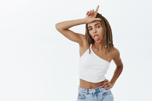 Sassy young african-american girl mocking someone, showing loser gesture on forehead and tongue