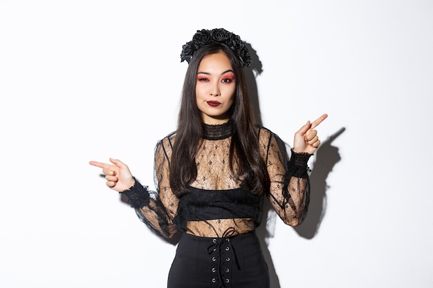 Free Photo sassy young evil witch with gothic makeup and wreath, looking arrogant while pointing fingers sideways, showing two halloween themed banners, standing over white background.