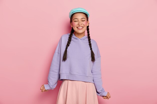 Satisfied adorable young Korean female model with rouge cheeks, laughs from joy, wears blue cap, oversized jumper and skirt