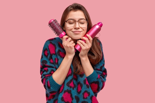 Free photo satisfied beautiful brunette lady has dreamy expression, carries hairbrush and spray for making coiffure, wears big eyewear