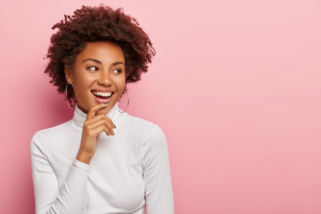 Satisfied carefree female model smiles gently, touches chin, looks aside, notices funny scene, laughs at something, has natural curly dark hair, dressed casually, isolated on pink wall