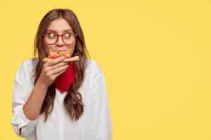 Free photo satisfied caucasian model eats delicious pizza indoor, has lunch, wears optical glasses, white shirt and red bandana, stands against yellow wall with free space for your slogan or text