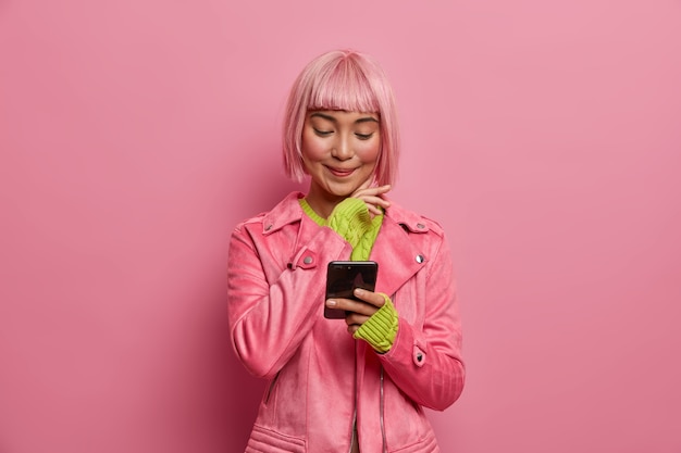 Free photo satisfied charming woman social media blogger with stylish hairstyle, holds smartphone, reads article in internet