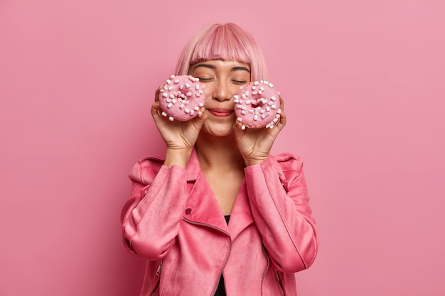 Satisfied charming woman with pink hair and fringe, closes eyes, imagines pleasant taste of donuts, dressed in rosy jacket