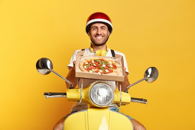 Free Photo satisfied deliveryman with helmet driving yellow scooter while holding pizza box