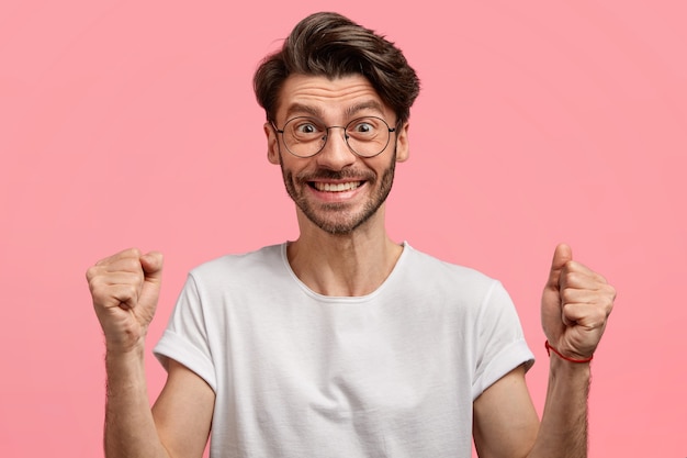 Free Photo satisfied emotive unshaven man with trendy haircut, thick stubble, clenches fists with success and victory, wears white t shirt and spectacles, isolated over pink space