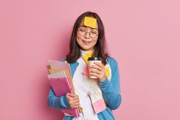 Satisfied female manager works with paper documents has coffee break closes eyes sticky note with drawn graphic stuck to forehead wears round spectacles.