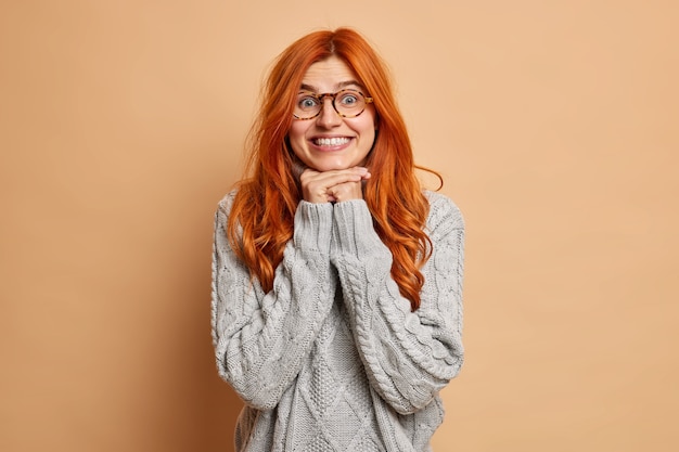 Satisfied female model keeps hands under chin looks with curious expression smiles broadly dressed in knitted sweater.