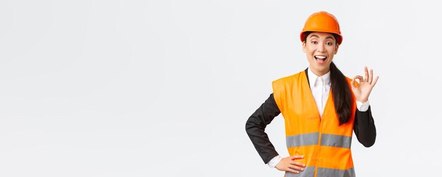 Satisfied happy asian female engineer construction manager in safety helmet and reflective jacket showing okay gesture pleased guarantee building quality ensure everything excellent