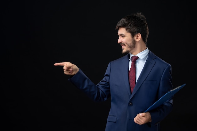 Satisfied male office worker in suit holding documents and pointing something on the right side on isolated dark wall