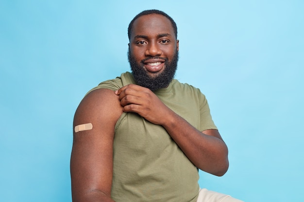 Satisfied man after vaccination shows adhesive bandage on arm got covid 19 vaccination to cure disease isolated over blue wall