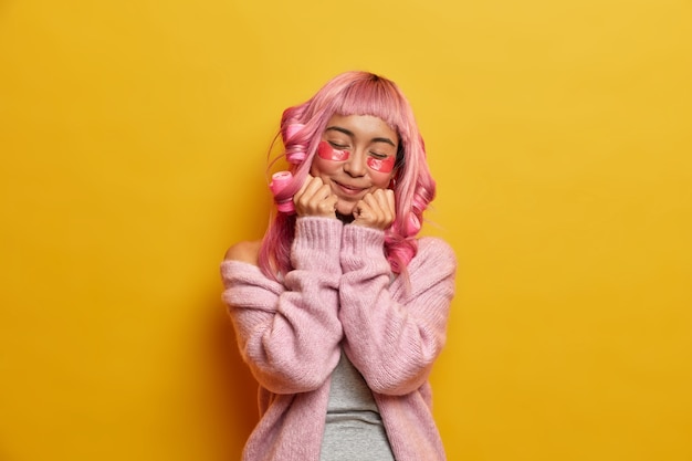 Free Photo satisfied pleased pink haired asian woman keeps hands under chin, closes eyes, applies hair rollers